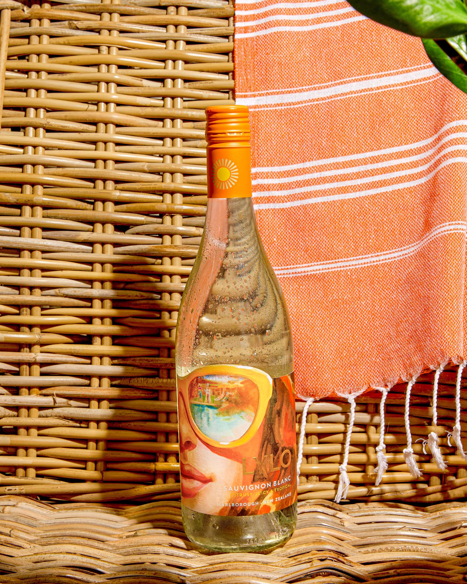 A bottle of Sauvignon Blanc with an orange label featuring sunglasses, set against a wicker background. An orange and white striped cloth is draped beside it, creating a summery vibe.