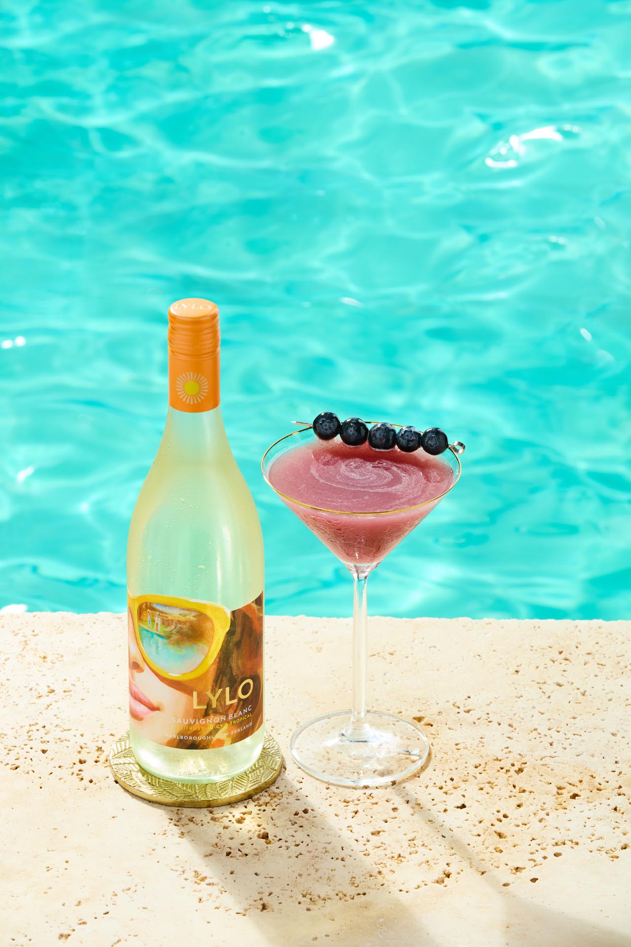 A bottle of Lylo wine and a martini glass filled with a pink cocktail topped with blueberries are on a poolside ledge. The turquoise water of the swimming pool sparkles in the background.