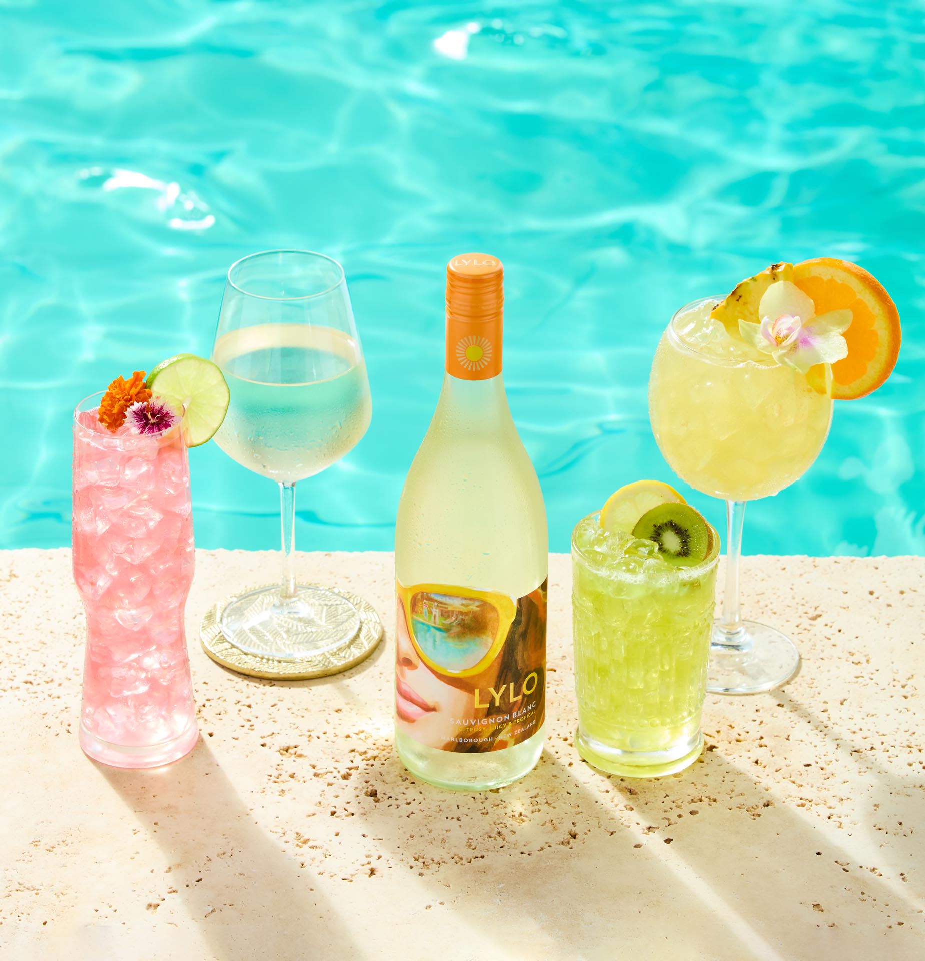 A bottle of Lylo wine by a pool, surrounded by four different cocktails: a pink drink with flowers and lime, a white wine in a glass, a yellow drink with an orange slice and flower, and a green drink with a kiwi slice.