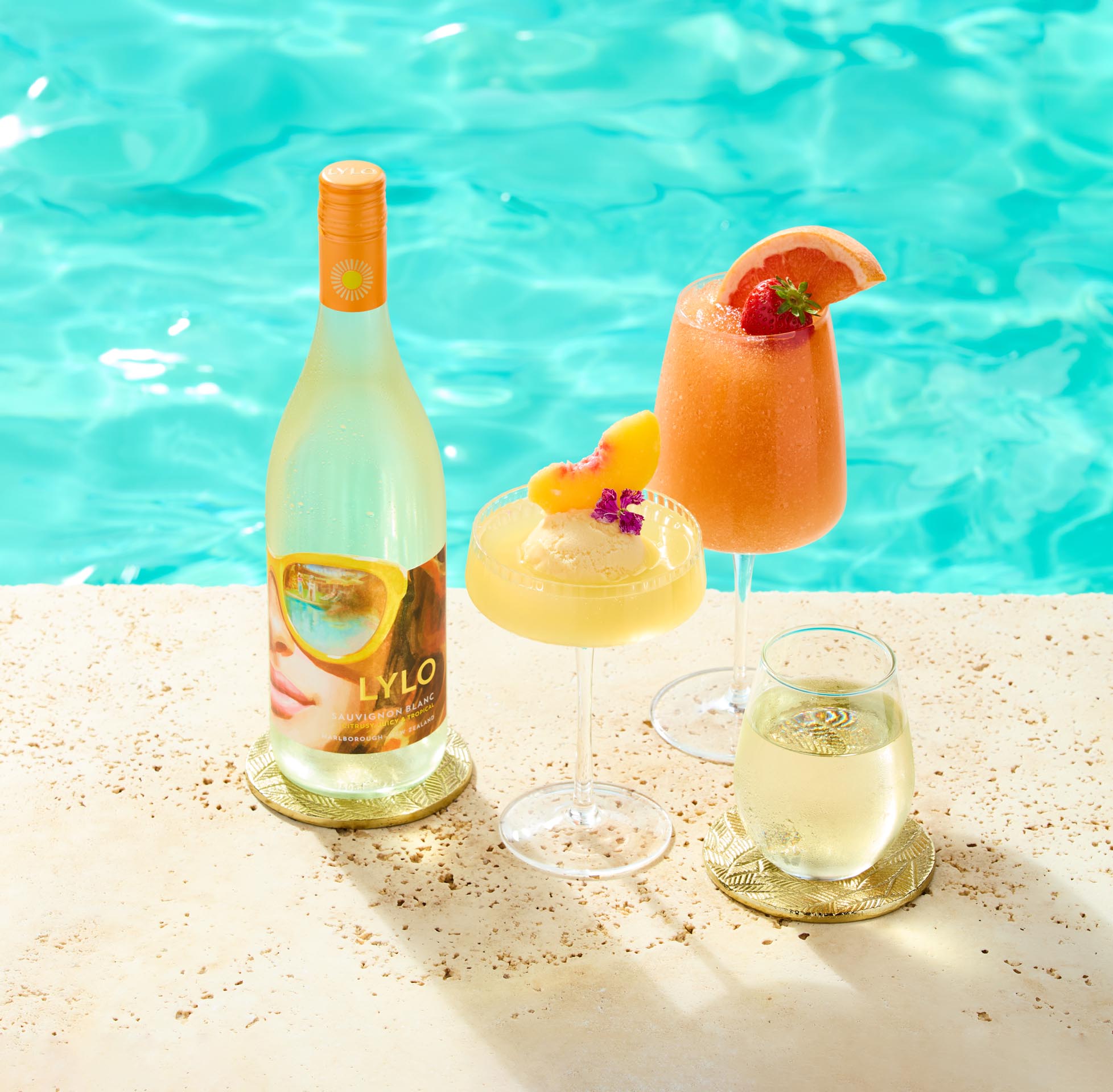 A refreshing poolside setup featuring a bottle of white wine, two wine glasses with chilled drinks garnished with fruit and flowers, and a third glass with wine. The backdrop is a clear turquoise pool, creating a sunny, tropical vibe.