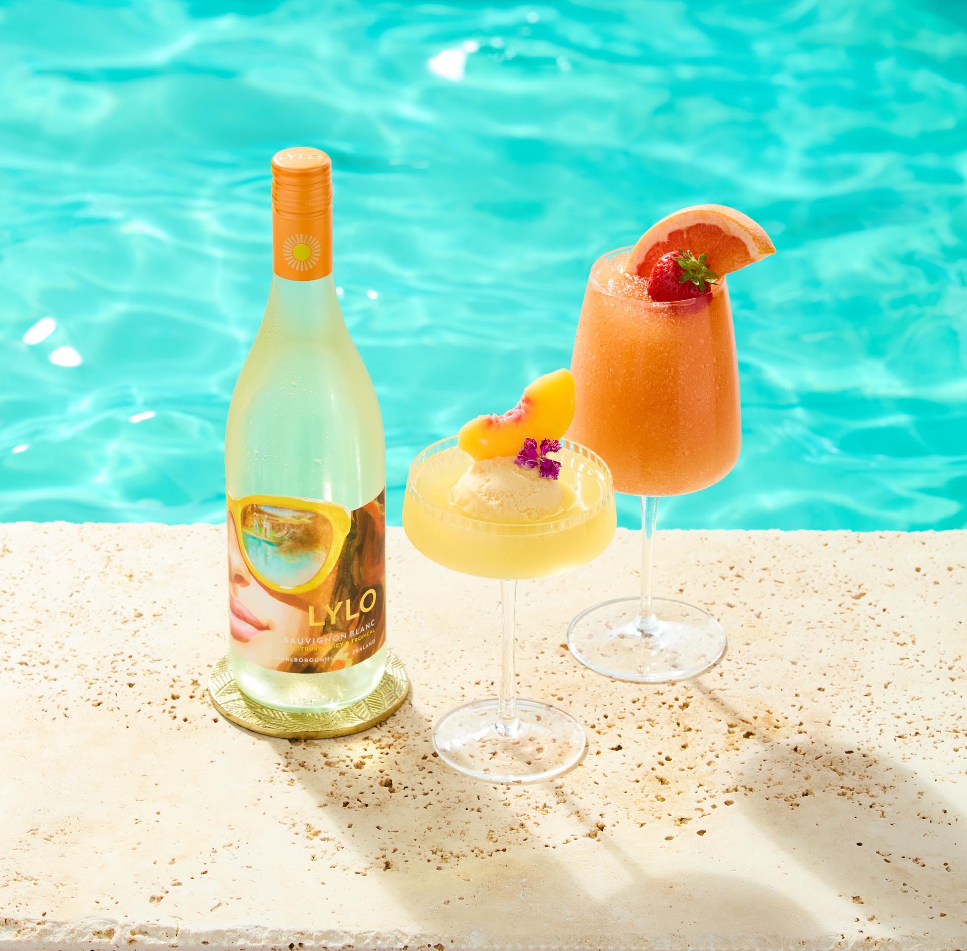 A refreshing poolside setup featuring a bottle of white wine, two wine glasses with chilled drinks garnished with fruit and flowers, and a third glass with wine. The backdrop is a clear turquoise pool, creating a sunny, tropical vibe.