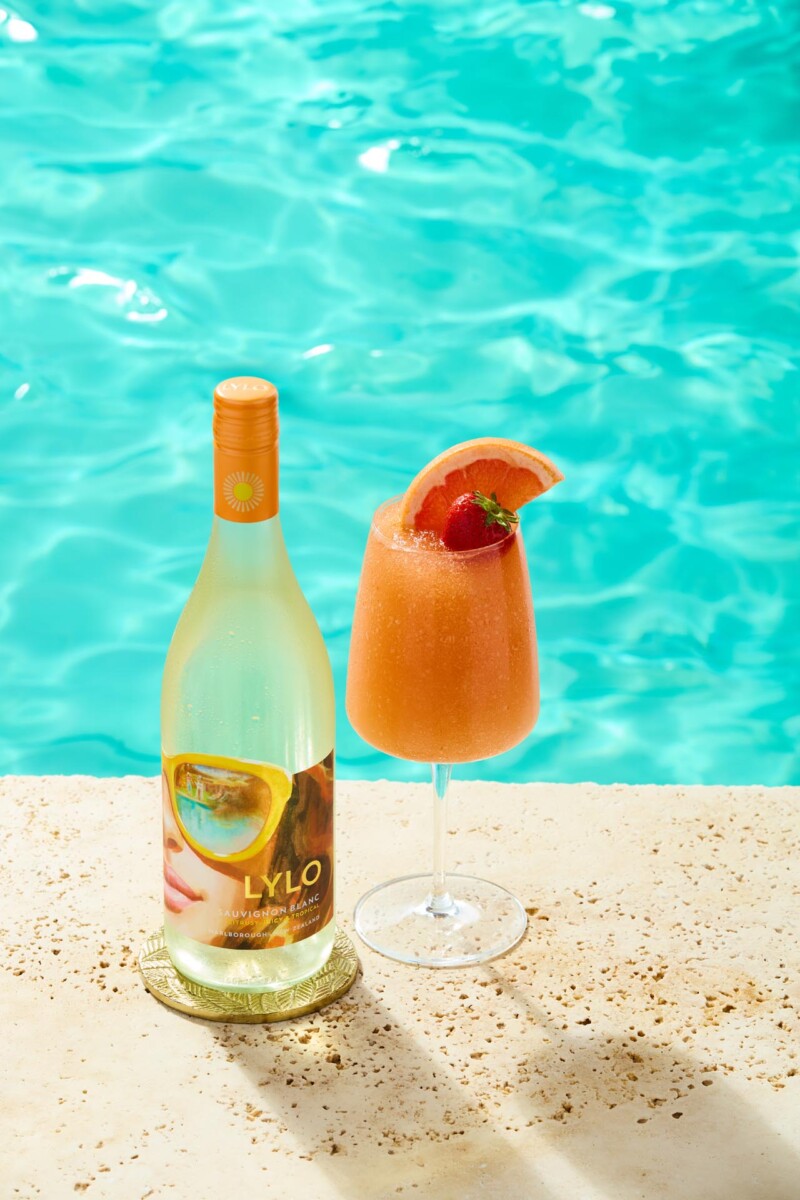 A bottle of LYLO grapefruit spritz stands on a poolside ledge next to a glass of orange slush garnished with a grapefruit slice and a strawberry. The clear turquoise pool water glistens in the background.