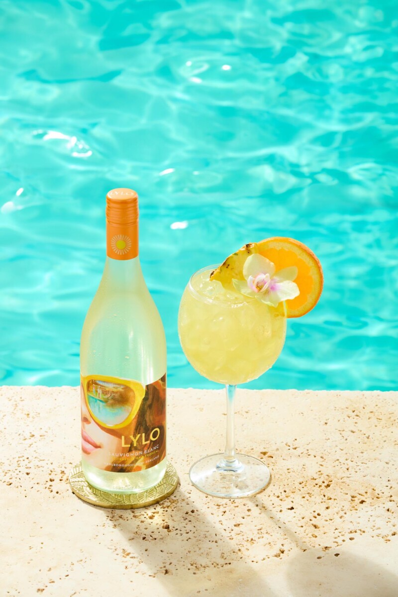 A bottle of LYLO tropical wine spritzer on a poolside surface next to a glass filled with a yellow drink, garnished with an orange slice, pineapple piece, and orchid flower. In the background, a turquoise pool is visible.