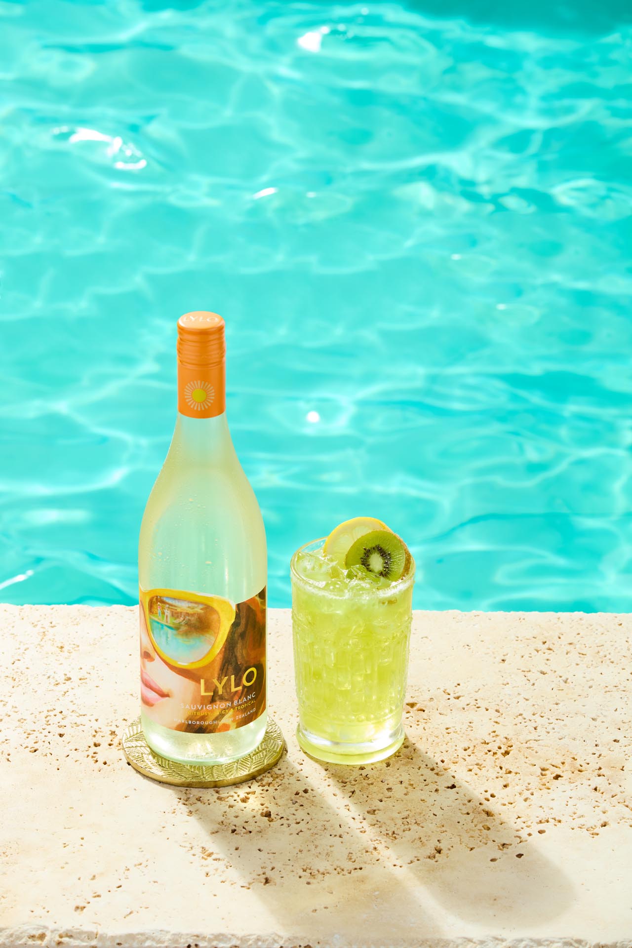 A bottle of Lylo wine and a glass of kiwi-flavored beverage sit on a poolside ledge. The glass is garnished with a kiwi slice. The clear blue water of the pool is visible in the background.