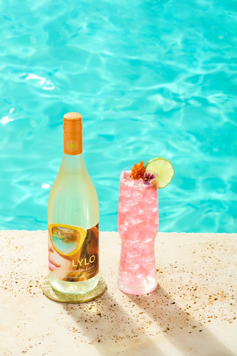 A bottle of Lylo Sauvignon Blanc stands next to a tall glass filled with pink cocktail, garnished with an orange twist, lime slice, and flower, by the edge of a swimming pool. The pool water is a bright turquoise.