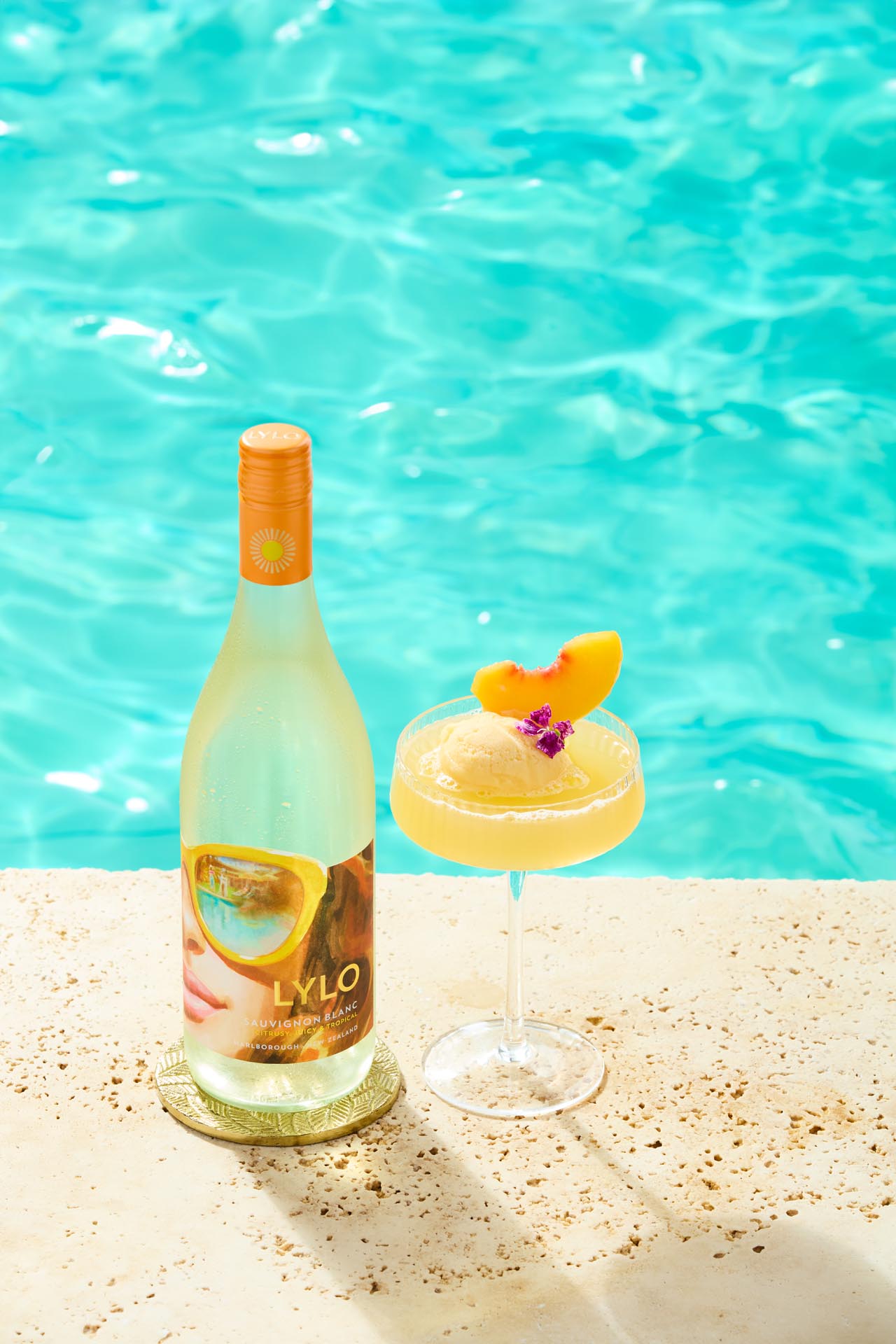 A bottle of Lylo wine is placed next to a cocktail glass with a peach garnish and a small purple flower, sitting on a textured stone surface by a shimmering turquoise pool.