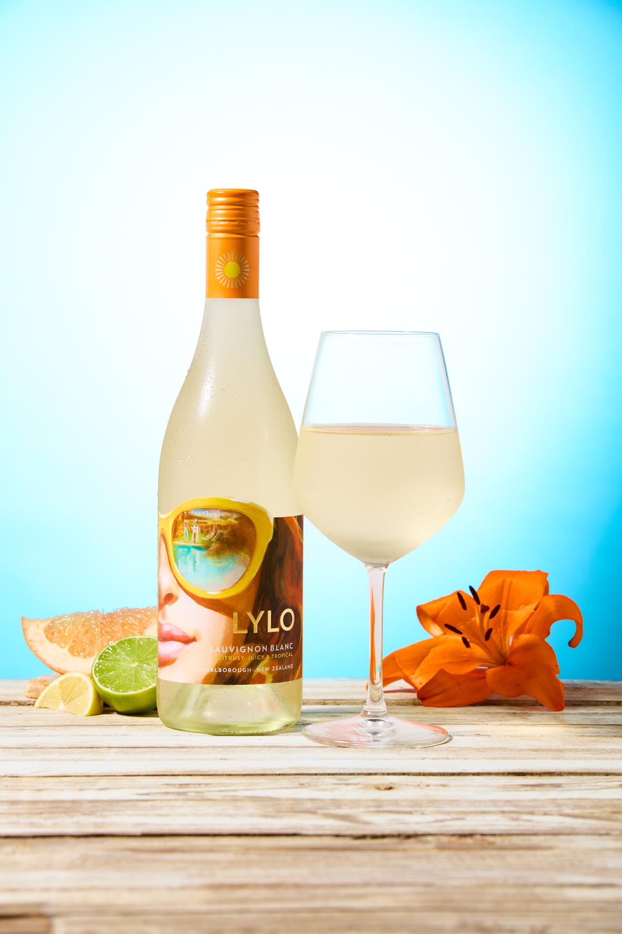 A bottle of Lylo Sauvignon Blanc with a sunny label, a glass of white wine, a slice of grapefruit, lime, lemon, and an orange lily on a wooden surface against a bright blue background.