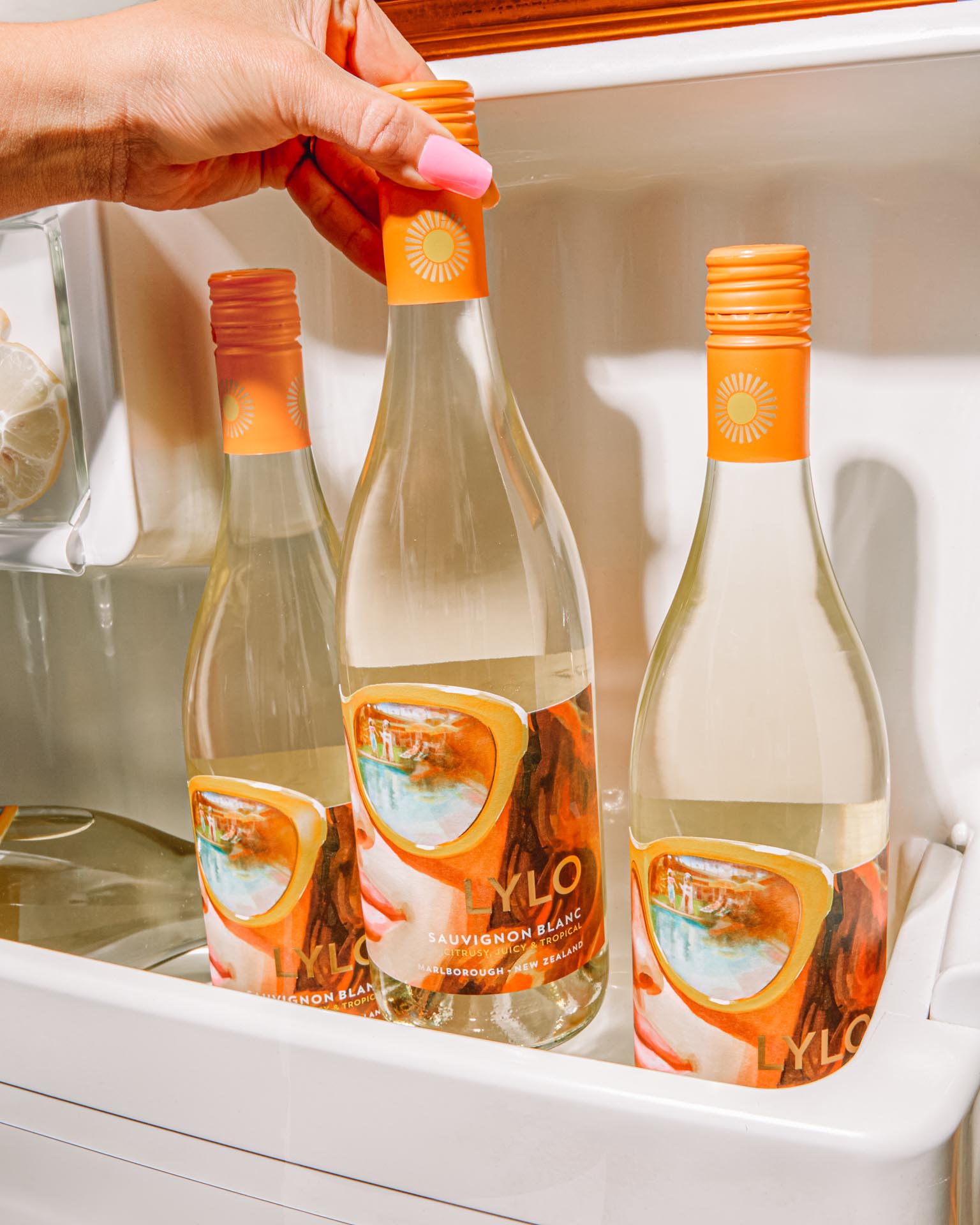 A hand is reaching into a refrigerator door shelf, selecting one of three bottles of LYLO Sauvignon Blanc. The bottles have orange caps and labels featuring a colorful design.