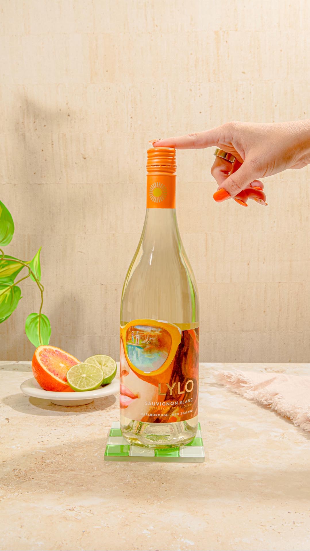 A hand with painted nails opens a bottle of Sauvignon Blanc on a table. The bottle has a colorful label. Nearby, there are sliced oranges and limes on a plate and a green plant in the background.