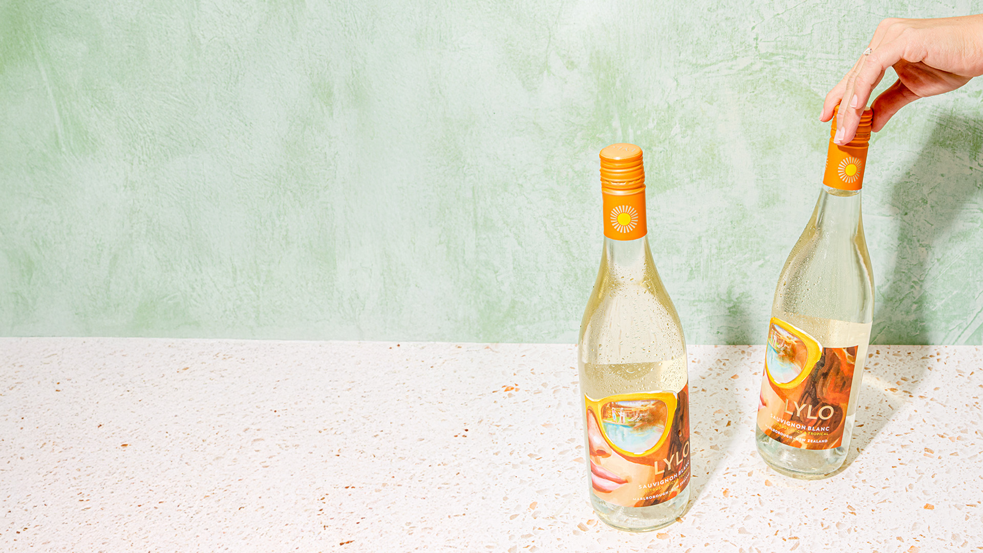 Two bottles of LYLO wine with orange caps are placed on a speckled white surface against a mint green background. A hand is touching the cap of one bottle.